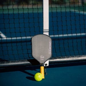 pickleball - Montana Contractors Association Padel racket leaning against a tennis net with a neon yellow-green ball on a blue court.